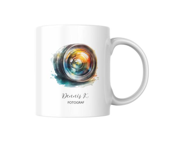 Eine weiße Keramiktasse mit einem farbenfrohen Motiv eines Kameraobjektivs. Das Objektiv ist in lebendigen Farben gestaltet, umgeben von aquarellierten Farbspritzern. Darunter steht der Name „Dennis K.“ in eleganter Schrift, gefolgt von dem Wort „FOTOGRAF